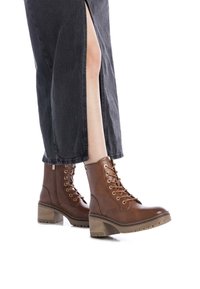 Botas de tobillo de cuero marrón con diseño de cordones, parte superior texturizada, tacón grueso y suela con relieve, combinadas con pantalones de pierna ancha negros con abertura lateral.