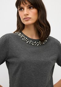 Šedý svetr s kulatým výstřihem, který má ozdobné uspořádání perel kolem límce pro zvýšení textury a vizuálního zájmu.