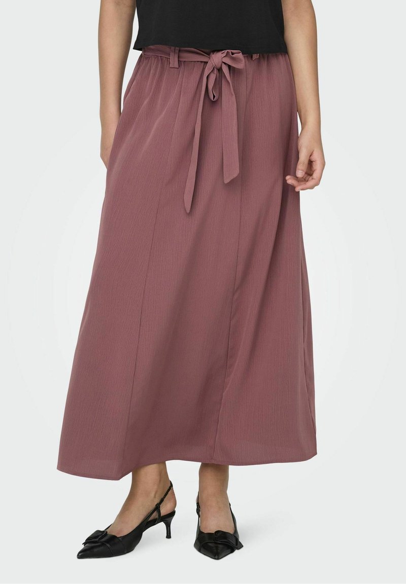 Femme portant une jupe midi mauve taille haute avec une ceinture nouée et des escarpins noirs à brides arrière à petit talon, debout devant un fond uni.