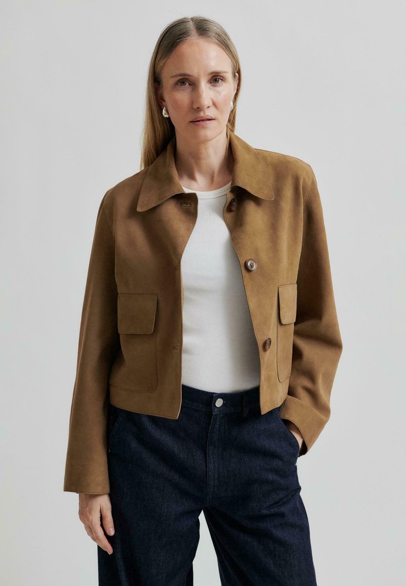 Giacca corta in suede marrone con colletto a punta, due tasche frontali e chiusura a bottone, indossata sopra una maglietta bianca e jeans scuri.