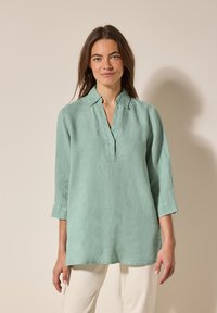 Camicia di lino verde chiaro con colletto corto, pattina dei bottoni e maniche a tre quarti. Il tessuto ha una texture morbida e una vestibilità rilassata.