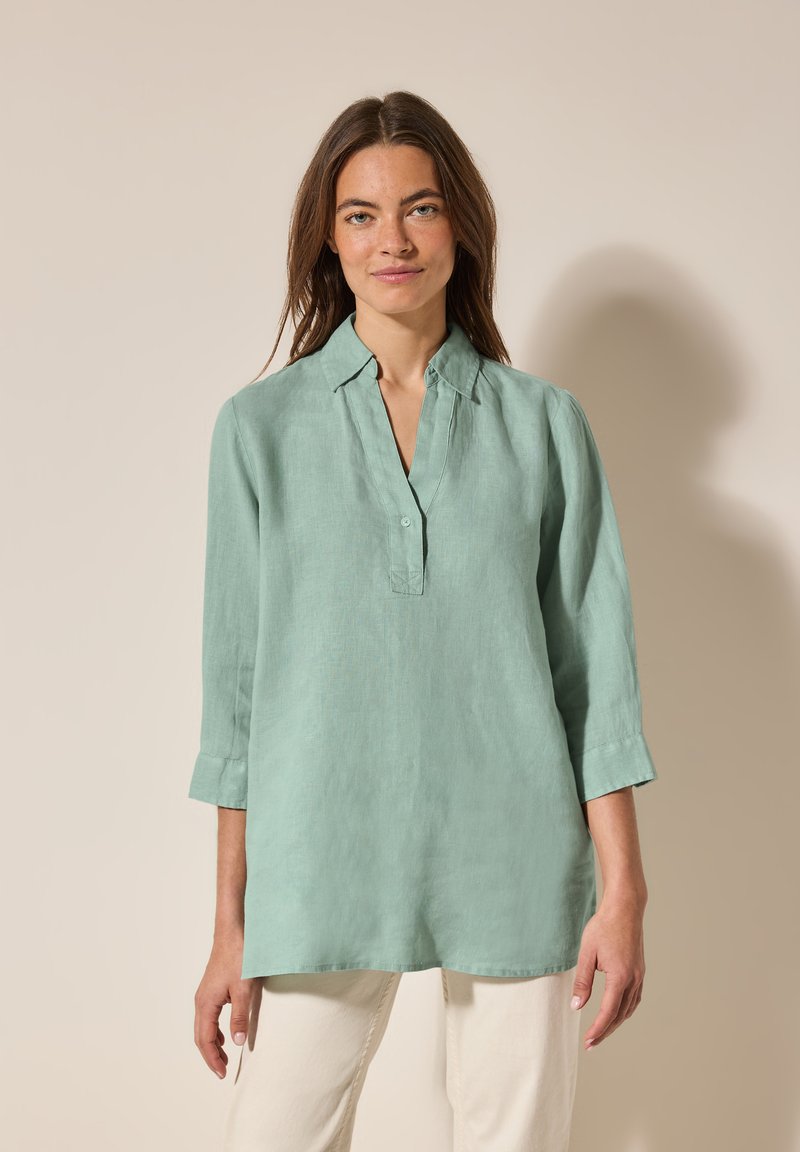 Camicia di lino verde chiaro con colletto corto, pattina dei bottoni e maniche a tre quarti. Il tessuto ha una texture morbida e una vestibilità rilassata.