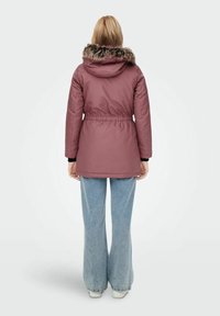 Veste isolante rose avec capuche en fausse fourrure, taille cintrée et manches longues ; portée avec un jean large bleu clair.
