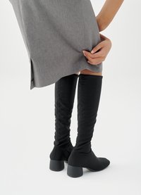 Μαύρες υφασμάτινες μπότες έως το γόνατο με εφαρμοστή γραμμή και τακούνι τετράγωνου σχήματος, συνδυασμένες με γκρι φούστα μεσαίου μήκους, που παρουσιάζει ομαλή υφή.