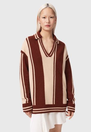 Jong persoon met lang, platinablond haar draagt een oversized bruin-beige gestreepte trui met V-hals over een witte rok.