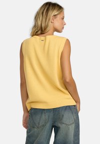 Top sans manches en tricot jaune avec un ourlet côtelé, doté d'une petite étiquette en métal au niveau du col, associé à un jean en denim ample.