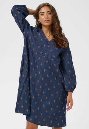 Donna con capelli ricci che indossa un vestito blu a maniche lunghe con piccoli motivi floreali marroni, posando con un braccio piegato dietro la testa.