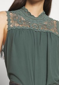 Top verde senza maniche con dettagli in pizzo intricati sullo scollo, texture del tessuto morbida, silhouette fluida e design arricciato sotto il petto.