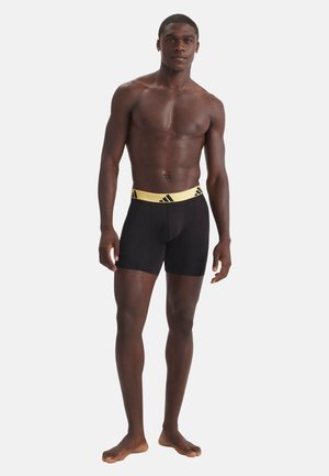 Atletisk mand stående barfodet iført sorte boksershorts med en gul linning, der har et gentaget sort logomønster.