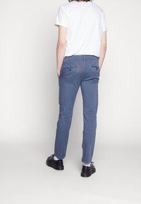 Modré úzké kalhoty s hladkou texturou, se dvěma zadními kapsami s knoflíkovými detaily. Kombinovány s bílým tričkem a černými botami.