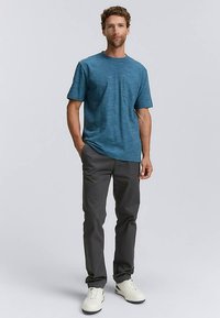 T-shirt bleu sarcelle à manches courtes avec une texture subtile, associée à un pantalon gris foncé et des baskets blanches. Design simple, coupe régulière, détail poche.