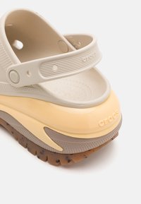 Beige Crocs med en matt overflate, gul glanset mellomsåle og strukturert brun yttersåle. Har en støttende justerbar stropp.