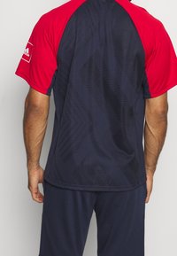 T-shirt de sport à manches courtes avec une partie supérieure rouge et une partie inférieure bleu marine. Présente des motifs texturés et un col rond. Logo Adidas sur la manche.