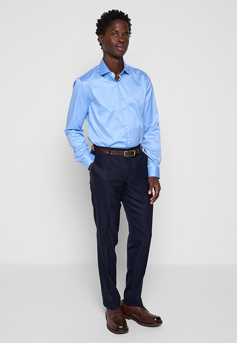 Hellesblaue Langarmbluse mit Knopfleiste; kombiniert mit dunklen Hosen und braunen Lederschuhen; glattes Material und taillierte Passform.