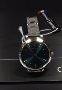 Orologio in acciaio inossidabile argentato con quadrante turchese rotondo, indicatori delle ore dorati e bracciale metalllico. L'etichetta mostra il nome del marchio Pierre Lannier.