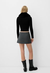 Kvinna klädd i en svart långärmad topp med pälskrage, grå mini kjol och svarta knälånga boots, stående vänd mot en vit vägg.