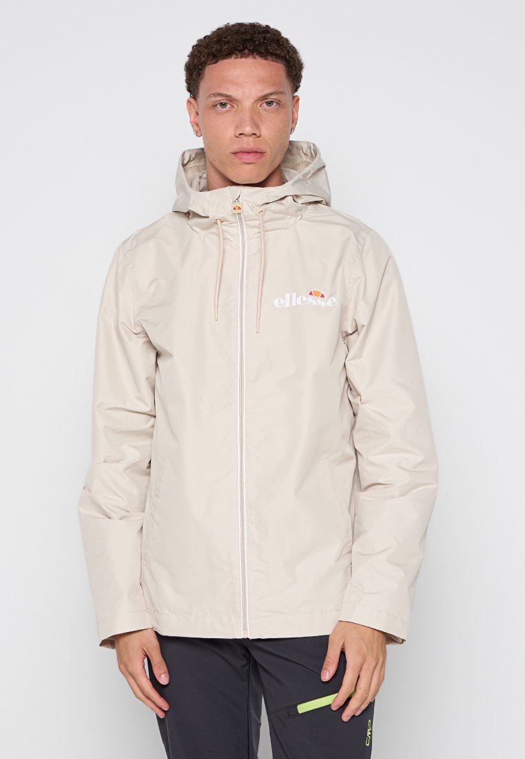 Chaqueta impermeable color beige claro con cierre frontal, capucha ajustable y puños elásticos. Presenta un logo en el pecho y bolsillos laterales.