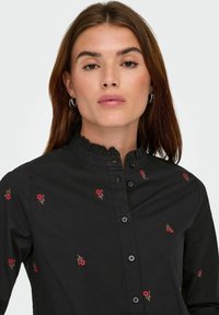 Giovane donna con capelli castani che indossa una camicia nera con bottoni, decorata con piccole ricami di fiori rossi e colletto a volant, su uno sfondo neutro.
