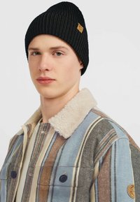 Sort ribbet beanie med et lille brunt logo-label, båret med en flerfarvet stribet jakke med en beige pelskrave og to knaplukninger.