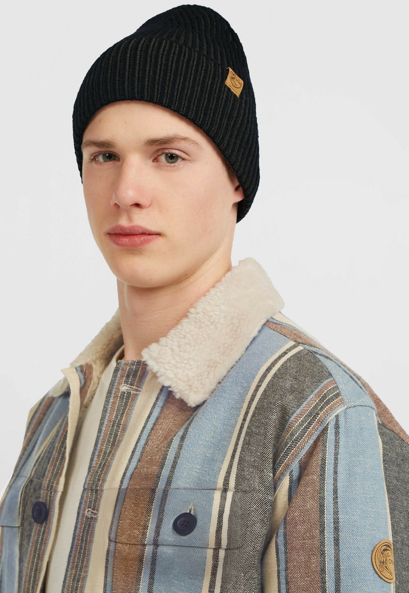 Sort ribbet beanie med et lille brunt logo-label, båret med en flerfarvet stribet jakke med en beige pelskrave og to knaplukninger.