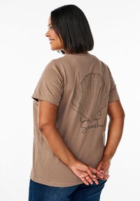Bruine katoenen t-shirt met een grote, gedetailleerde afbeelding van een zeehol en het woord "Seashell" in een donkere, handgeschreven lettertype op de achterkant.