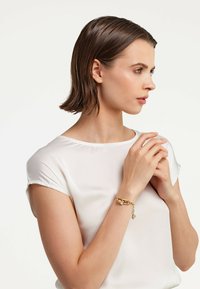 Auksinė grandinėlė su apvaliais akcentais, nešiojama ant riešo. Modelis vilki baltą, trumpasleeve viršutinį drabužį, turi tiesias vidutinio ilgio rudus plaukus.