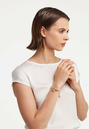 Goldene Kettenarmband mit runden Akzenten, getragen an einem Handgelenk. Das Modell trägt ein weißes, kurzärmliges Oberteil und hat glattes, mittellanges, braunes Haar.