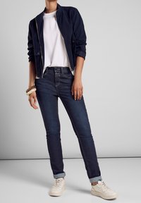 Marinblå bomberjacka, vit t-shirt, mörkblå skinny jeans med uppvikta mudd, och krämfärgade sneakers. Slät tyg med subtila sömmar.