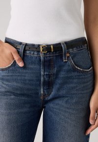 Μαύρη δερμάτινη ζώνη με χρυσή μεταλλική αγκράφα, φορεμένη πάνω από μπλε τζιν παντελόνι ψηλόμεσο με πορτοκαλί ραφές και φθαρμένες άκρες.