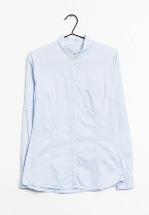 Chemise bleu clair à manches longues avec col et poche poitrine, suspendue sur un cintre noir sur fond blanc.