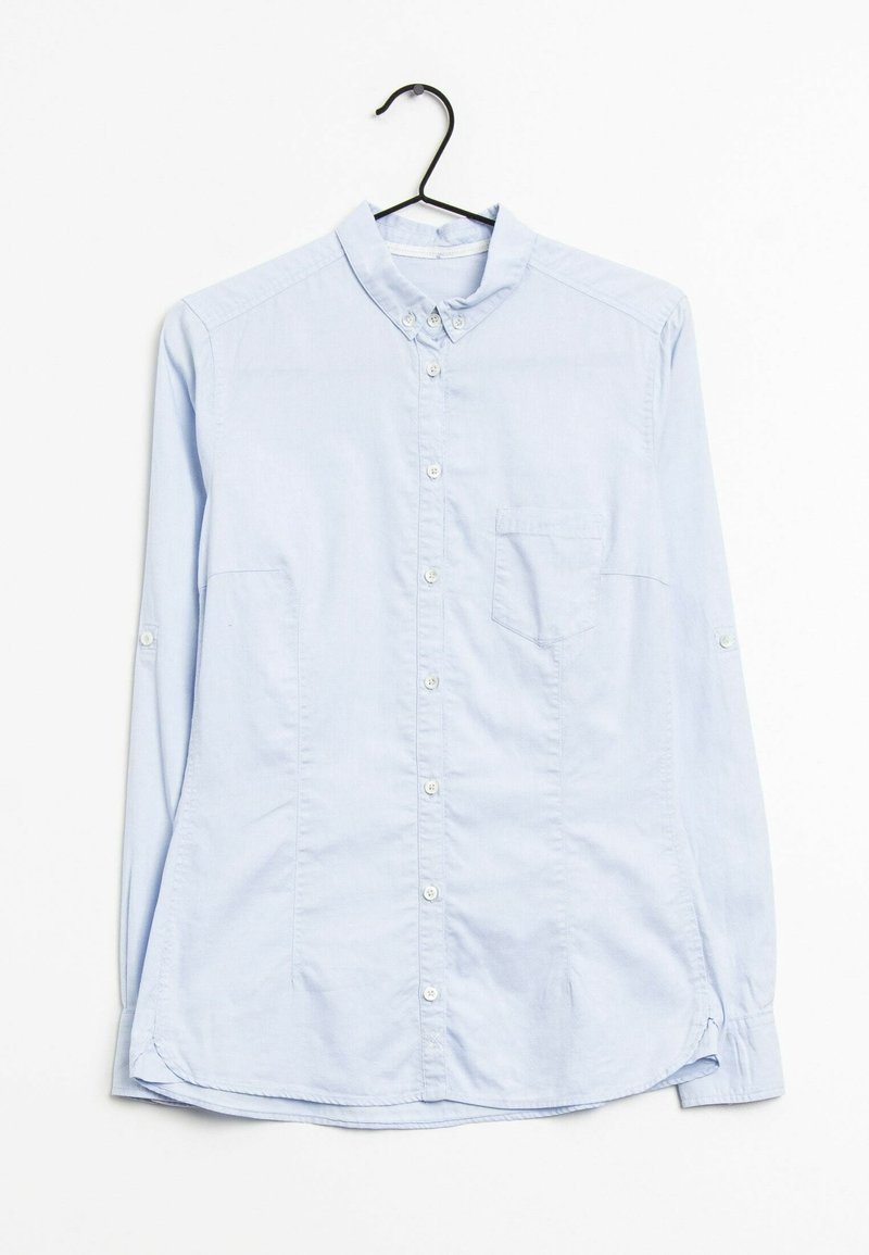 Chemise bleu clair à manches longues avec col et poche poitrine, suspendue sur un cintre noir sur fond blanc.