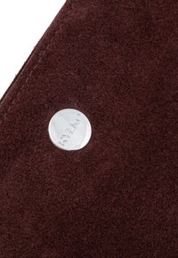 Bordo zomšo medžiaga su subtiliu tekstūru, su sidabriniu metalo logotipo akcentu. Nuotrauka užfiksuoja audinio ir detalių artimą planą.