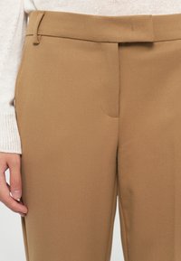 Pantalons beige sur mesure en tissu texturé, avec un devant plat, des passants de ceinture et des poches latérales. Gros plan mettant en valeur les détails de coupe.