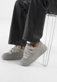 Helle graue Sneaker mit dicken strukturierten Schnürsenkeln, einem schlanken Design und einer robusten Sohle, kombiniert mit schwarzen Hosen, auf einer weißen Fläche platziert.