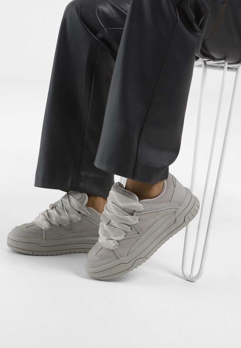 Helle graue Sneaker mit dicken strukturierten Schnürsenkeln, einem schlanken Design und einer robusten Sohle, kombiniert mit schwarzen Hosen, auf einer weißen Fläche platziert.