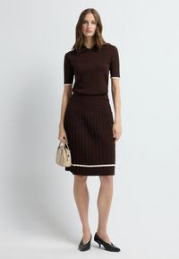Bruine gebreide jurk met geribbelde textuur, witte accenten op kraag en zoom, gecombineerd met zwarte hakken en een beige handtas.