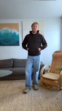 Black hooded sweatshirt, blue denim jeans, and gray shoes. Standing against a light wall with abstract paintings and a rattan chair nearby.