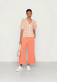 Blusa floral com mangas bufantes em tons de rosa e laranja, combinada com calças largas corais e ténis brancos. Acessórios incluem uma mala rosa claro.