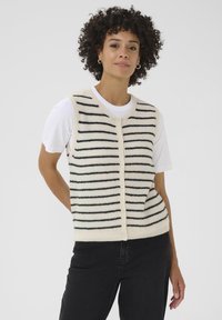 Donna sorridente con capelli ricci indossa un gilet a righe sopra una camicia bianca, posando di fronte a uno sfondo semplice. Mani nelle tasche, stile casual.