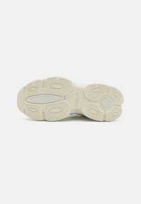 Sole di sneaker di colore chiaro con motivi di grip in gomma testurizzata, che presenta una sezione centrale liscia e un logo impresso di lato.