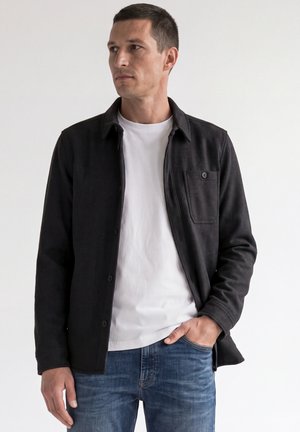 Mand iført en sort knappet jakke over en hvid t-shirt og blå jeans, der kigger til venstre med den ene hånd i lommen.