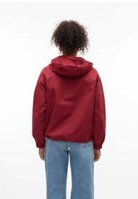 Personne aux cheveux bouclés portant une veste à capuche rouge et un jean bleu, vue de dos sur un fond blanc uni.