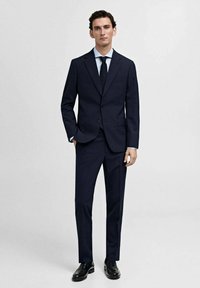 Marineblauw colbert-jack met een slim fit. Heeft een enkel-breasted blazer, notch revers en op maat gemaakte broeken. Gecombineerd met een lichtblauw overhemd en stropdas.