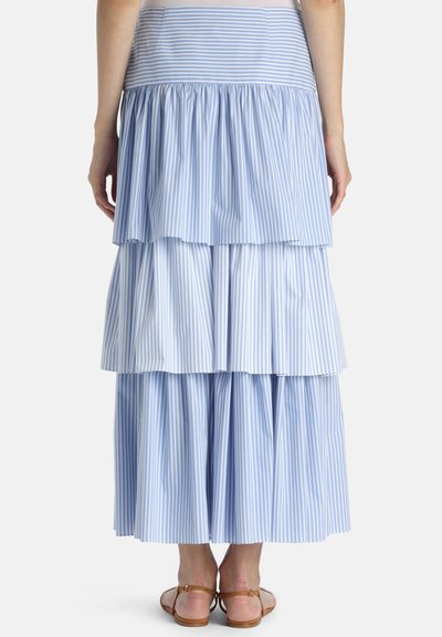 Kvinde iført en blå og hvid lodret stribet lagdelt nederdel og brune flade sandaler, vist bagfra mod en ensfarvet baggrund.