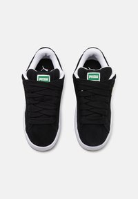 Baskets en suède noires avec semelles blanches, dotées d'une languette rembourrée, de lacets noirs et d'un logo Puma vert sur la languette.