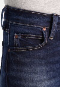 Pantalones vaqueros de mezclilla azul oscuro con un diseño clásico, que presentan un bolsillo frontal, costuras en contraste y una imagen en primer plano de la cinturilla.
