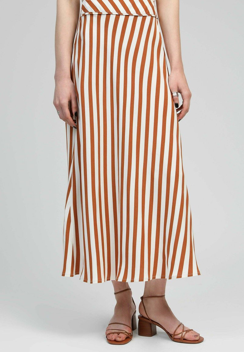 Jupe maxi rayée en blanc et rouille. Design fluide avec une large ceinture. Associée à des sandales à talons marron. Texture de tissu lisse.