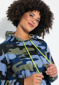 Felpa con cappuccio caratterizzata da un motivo camouflage blu, verde e nero. Presenta lacci in neon giallo e strass decorativi sul davanti.