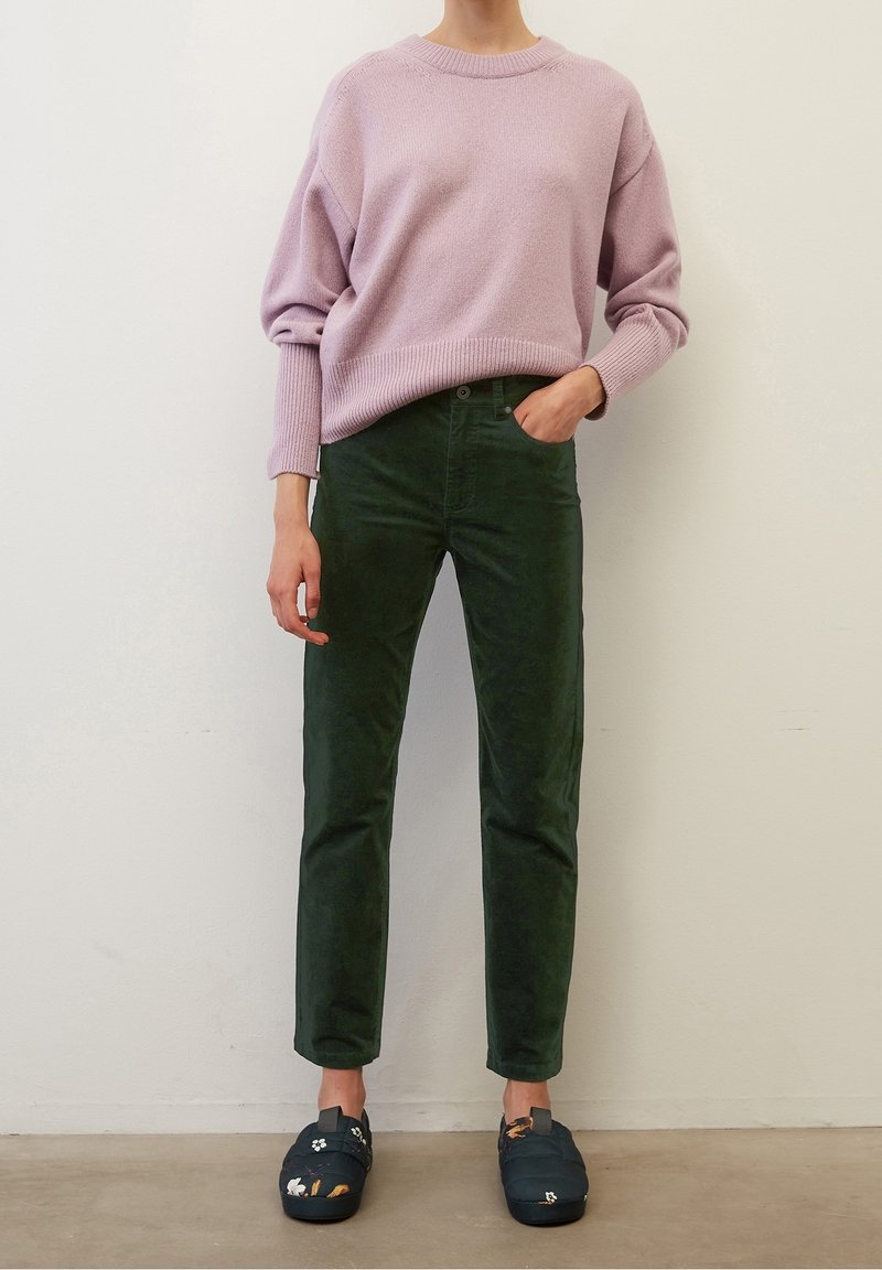 Femme portant un pull lilas, un pantalon en velours côtelé vert foncé et des chaussures slip-on noires à motifs floraux, debout contre un mur uni.