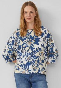 Haut à motif floral en bleu et beige, avec de larges manches 3/4, une coupe décontractée et un tissu texturé. Associé à un jean bleu.